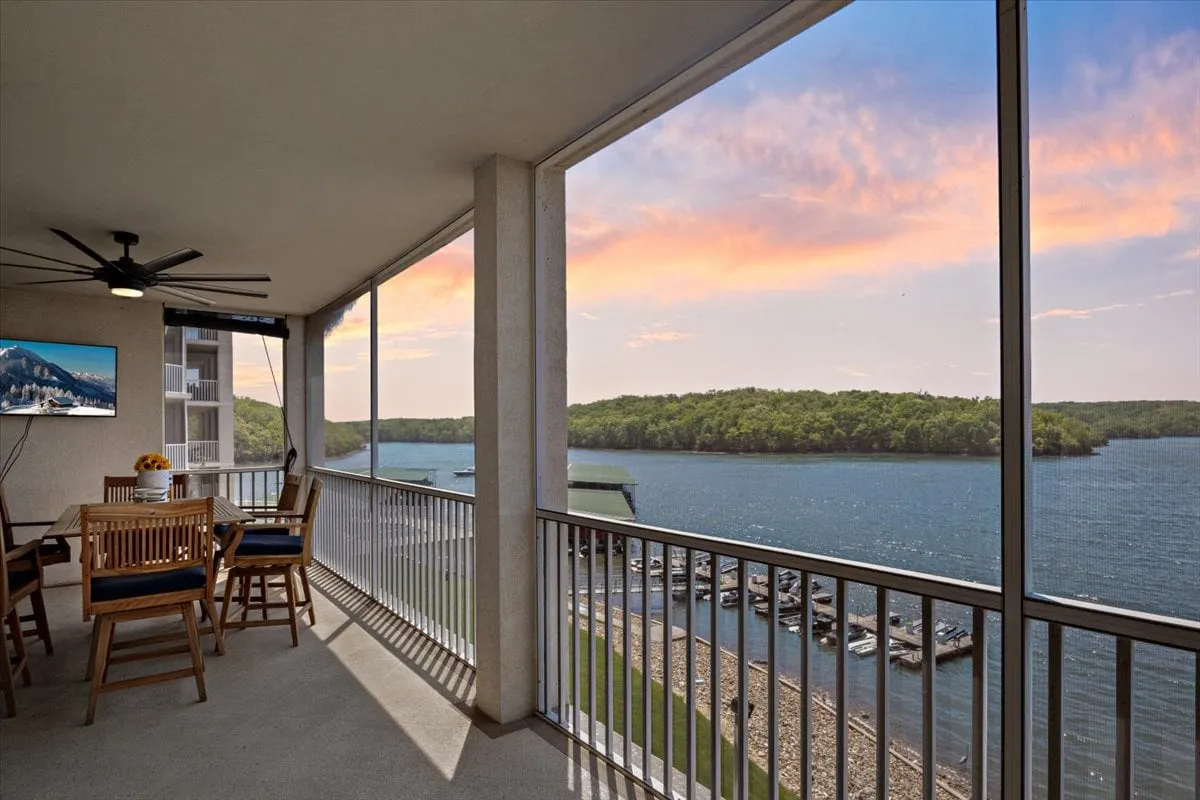 A private patio with a dining table and chairs, overlooking a scenic lake and a glowing sunset sky. The ceiling fan and inviting setup suggest comfortable outdoor dining and relaxation with direct access from the main living space.
