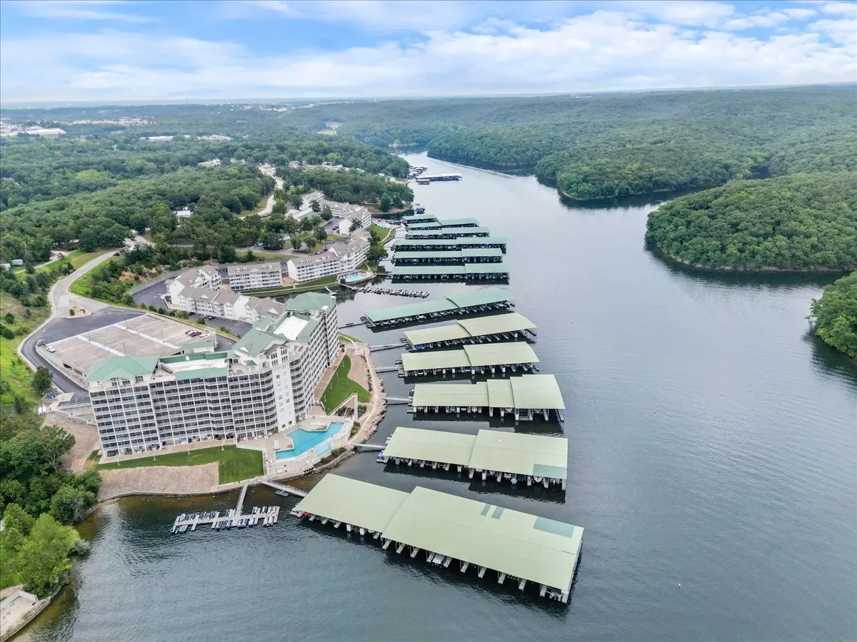 A wide exterior scene showing a large hotel building adjacent to a marina with boat docks, surrounded by lush green trees and calm water. The area is serene and clearly oriented towards lake activities such as boating, fishing, and enjoying sunsets by the water.