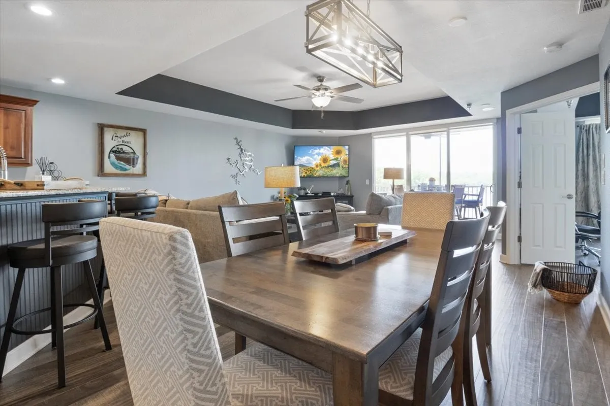 This image captures a spacious open-concept dining and living area, featuring a dining table, a bar area, a television, comfortable seating, and expansive windows that offer scenic views. The natural light streaming in highlights the inviting and connected atmosphere ideal for gatherings.