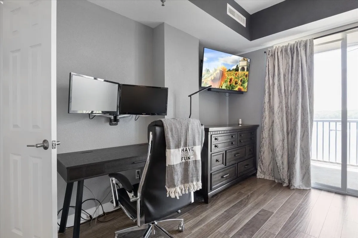 A work-ready bedroom corner with a dual monitor setup on a dark wooden dresser, accented by a large window with curtains. The room exudes productivity and comfort, blending a practical workstation with inviting bedroom touches.