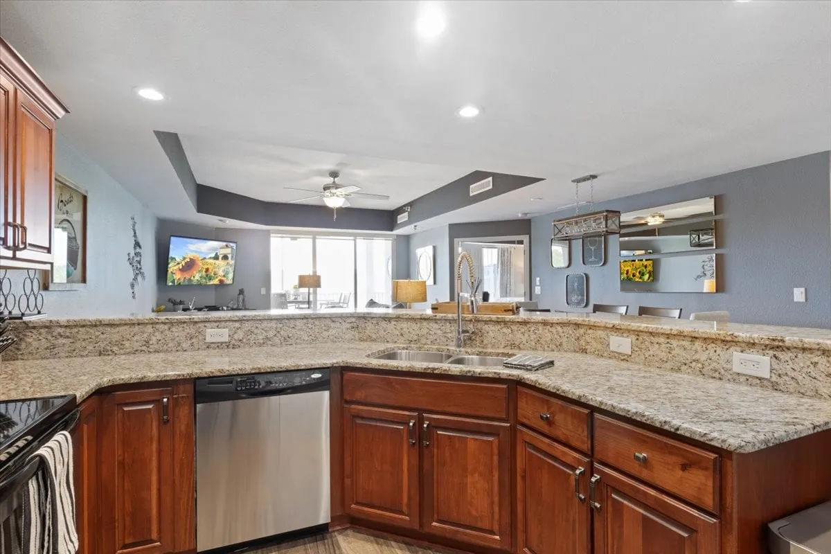 A modern kitchen with a granite countertop island, stainless steel appliances, ceiling fan, and large windows letting in abundant light. The kitchen design encourages social interaction, whether cooking or enjoying casual meals.