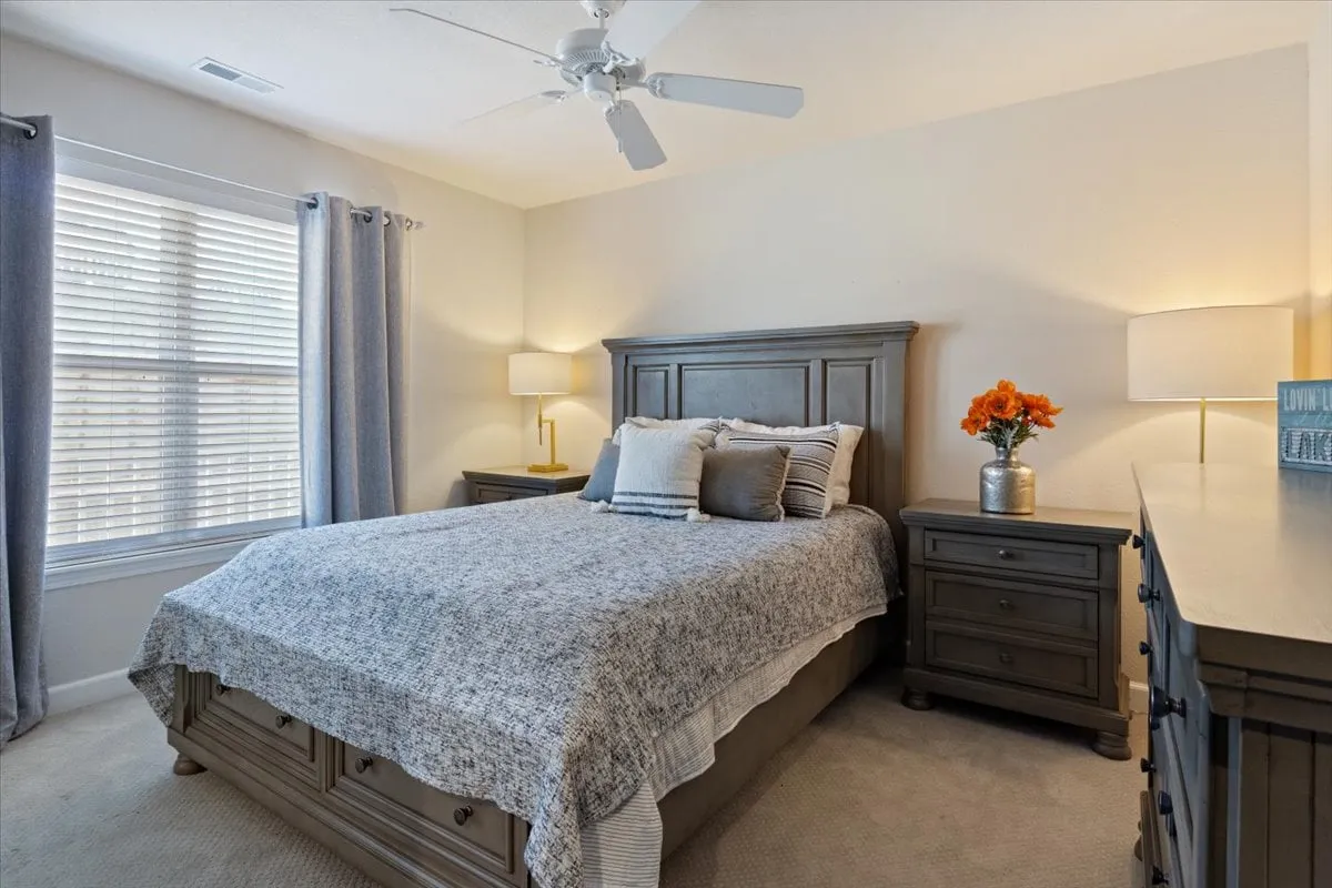 A serene bedroom scene with a cozy bed dressed in soft linens, flanked by nightstands and topped with a flower vase. Light pours in through a window, creating a tranquil, inviting space.