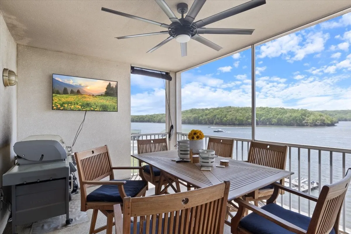 An inviting outdoor dining area with a table set for gatherings, bright sunflowers, a TV for entertainment, and an uninterrupted view of the water beyond. The space is clearly designed for enjoying meals and relaxing while soaking in the lakefront scenery.