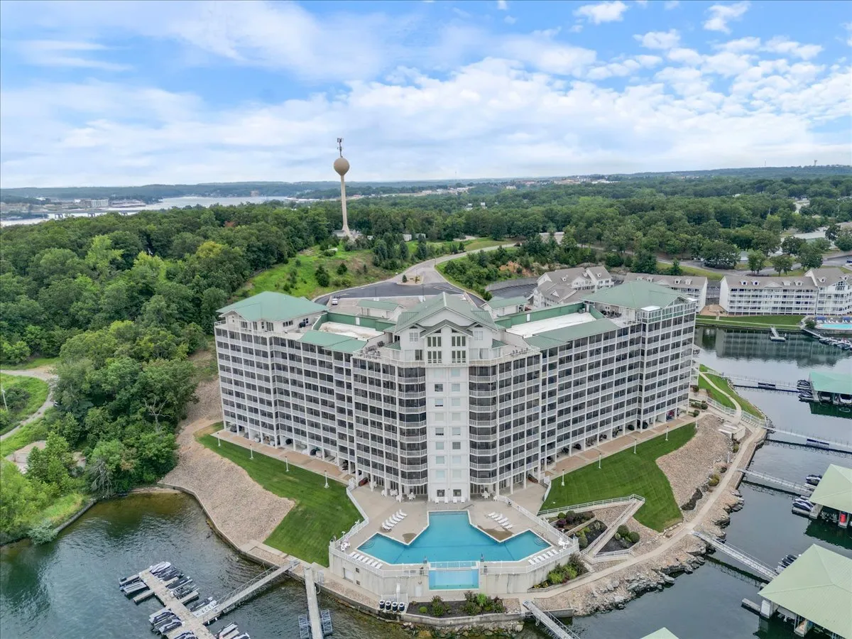 A broad exterior shot featuring the hotel building, a sparkling swimming pool, lush greenery, and an adjacent marina with boats. The scene is lively yet relaxing, with clear sightlines to the lake, inviting guests to imagine themselves swimming, boating, or lounging in this resort-like atmosphere.
