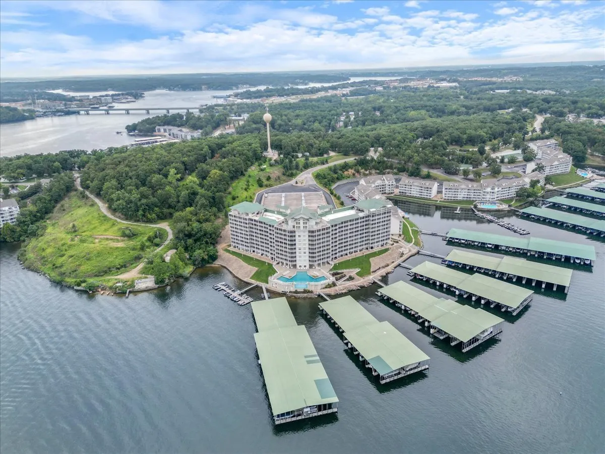 A sweeping exterior view of a large hotel building nestled beside a marina with boat slips, surrounded by lush greenery. The marina is clearly visible, with several boats moored at the docks, set against the backdrop of water and a water tower. Roads and bridges connect the property to the surrounding area, reinforcing its prime lakefront location.