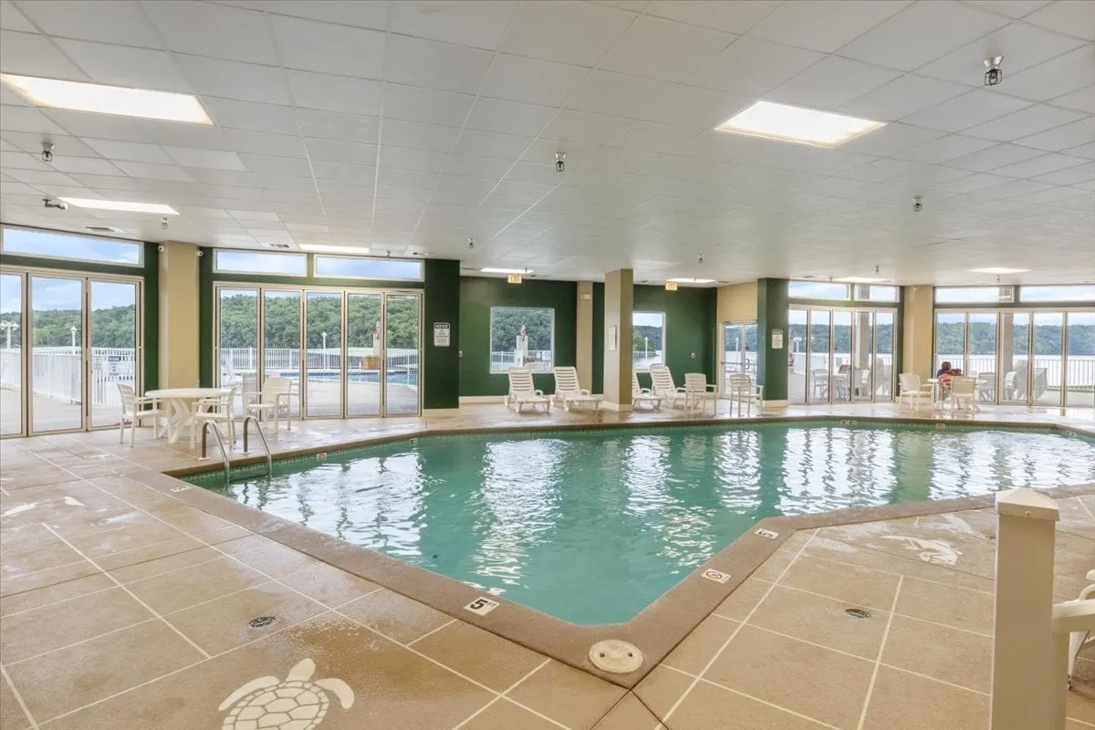 This image features a spacious indoor swimming pool area with large windows that allow natural light to flood in. Poolside chairs are arranged along one side, creating a tranquil and refreshing setting ideal for swimming, relaxing, or socializing in a climate-controlled environment.